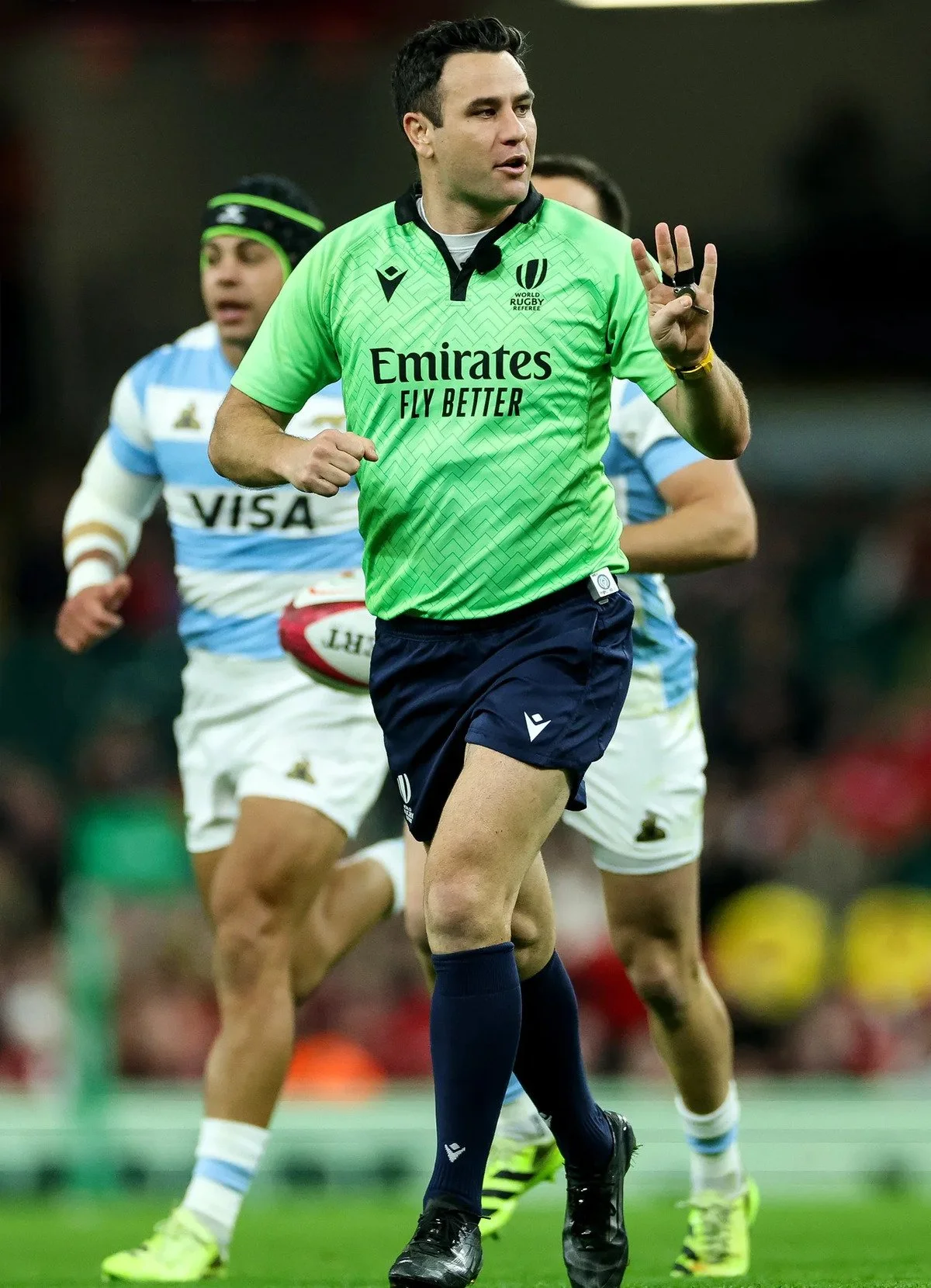 A rugby referee Ben O'Keeffe in a green jersey and dark blue shorts running on the field during a match, with rugby players in blue and white uniforms behind him.