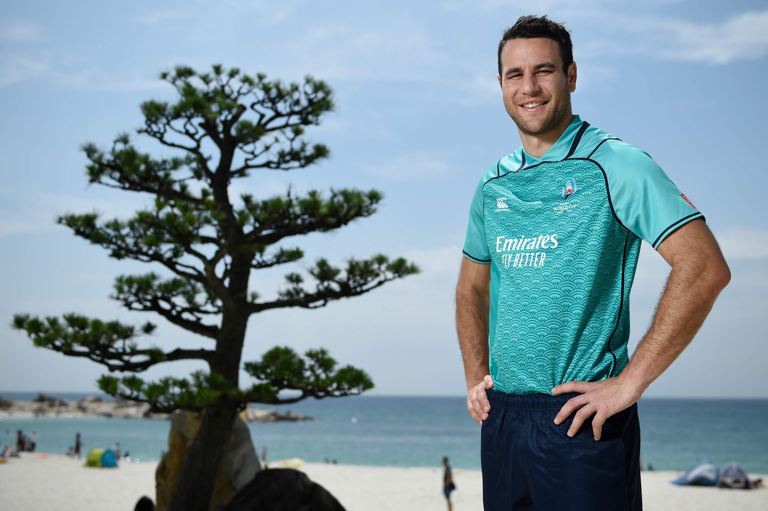 Ben O'Keeffe referee smiling in an aqua sports jersey stands on a beach with a tree, ocean, and people in the background.