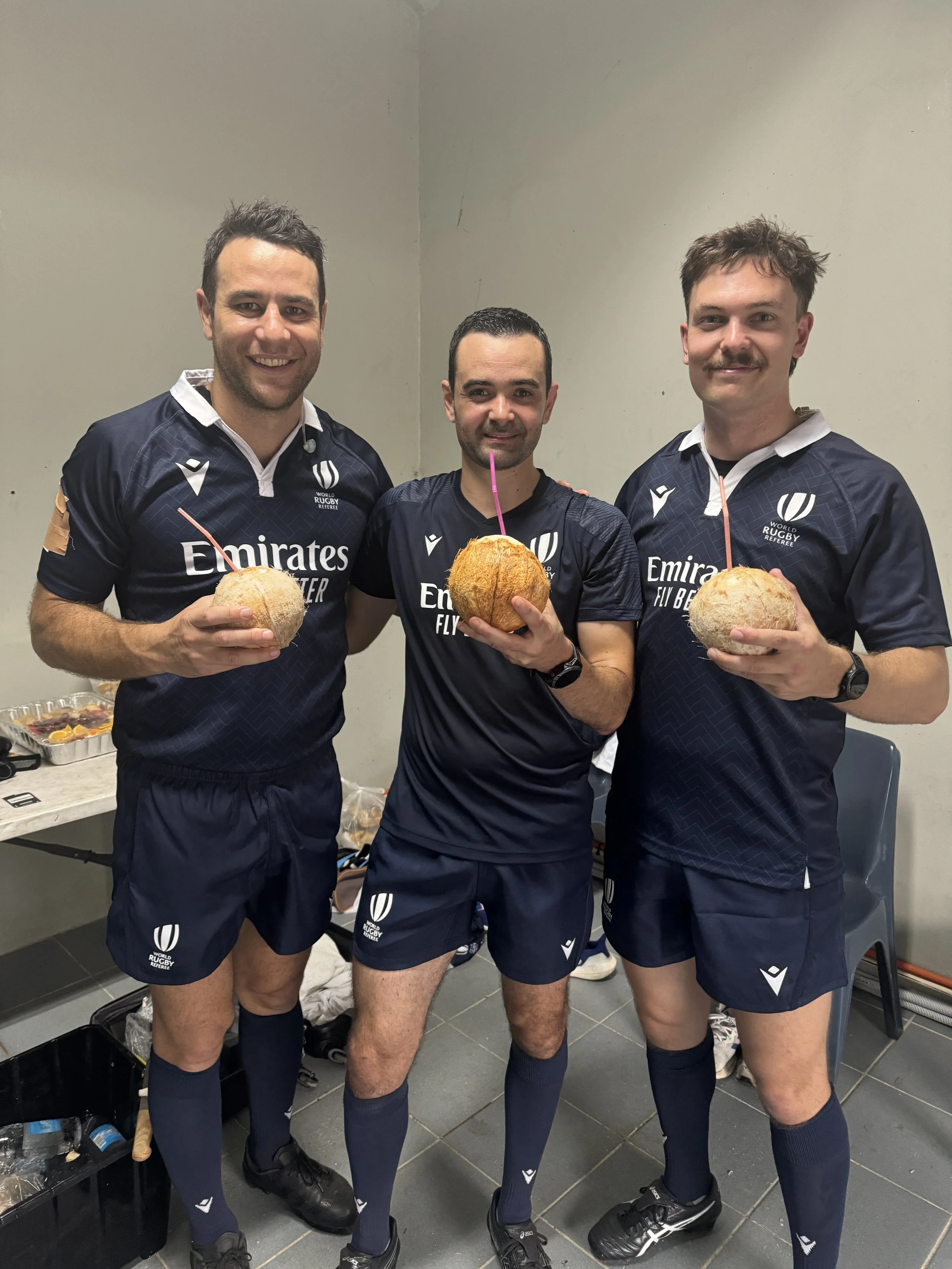 Three rugby referees, including Ben O'Keeffe, wearing navy blue jerseys and shorts, holding coconuts with straws, inside a room with a grey wall and grey tiled floor.