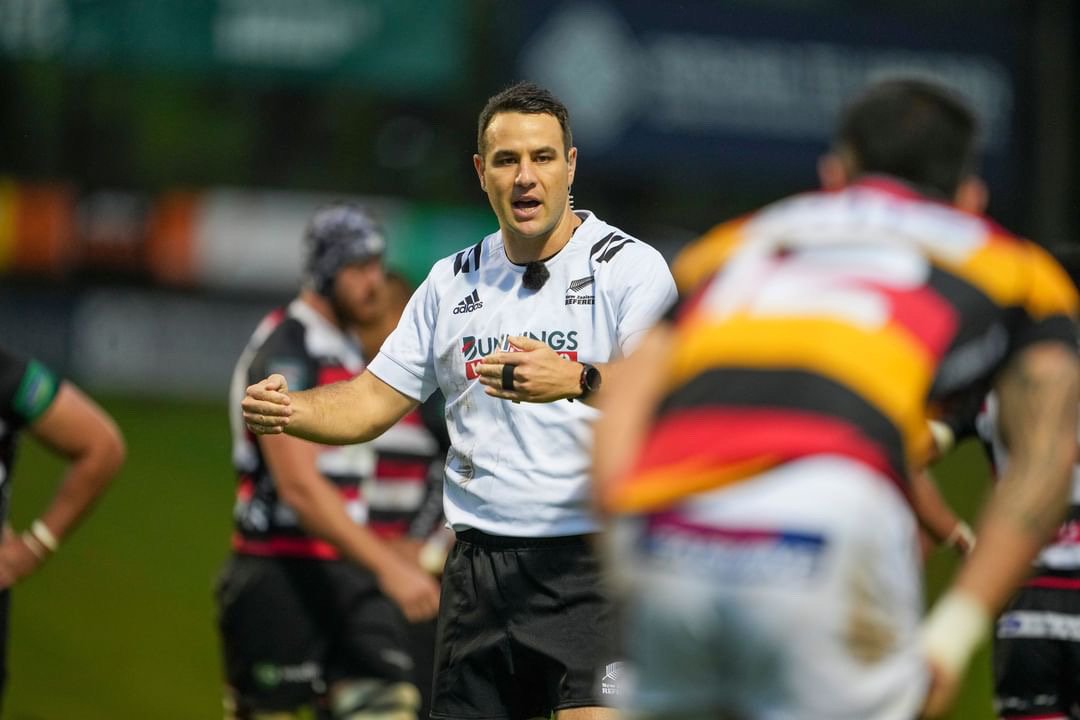 Rugby referee Ben O'Keeffe, wearing a white jersey with sponsoring logos, is talking to players on the field during a game, with teammates in black and red striped jerseys and an opposing team in yellow, black, and red jerseys in the background.