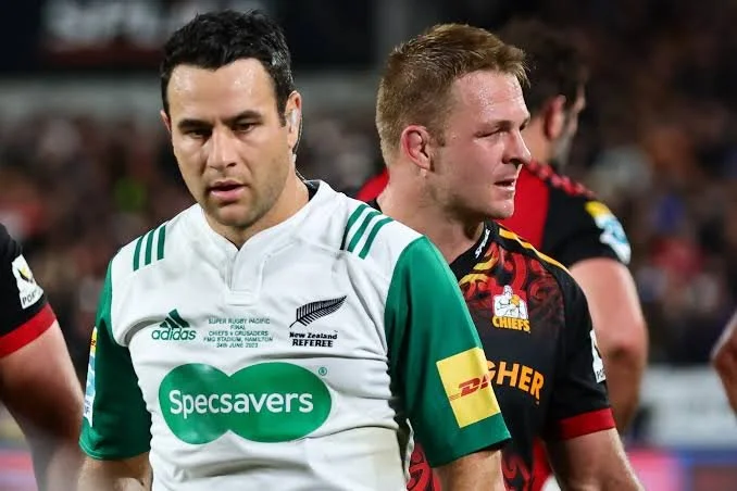 Two rugby players standing on the field, one in a green and white jersey is the referee Ben O'Keeffe and the other in a black and red jersey, with a blurred crowd in the background.