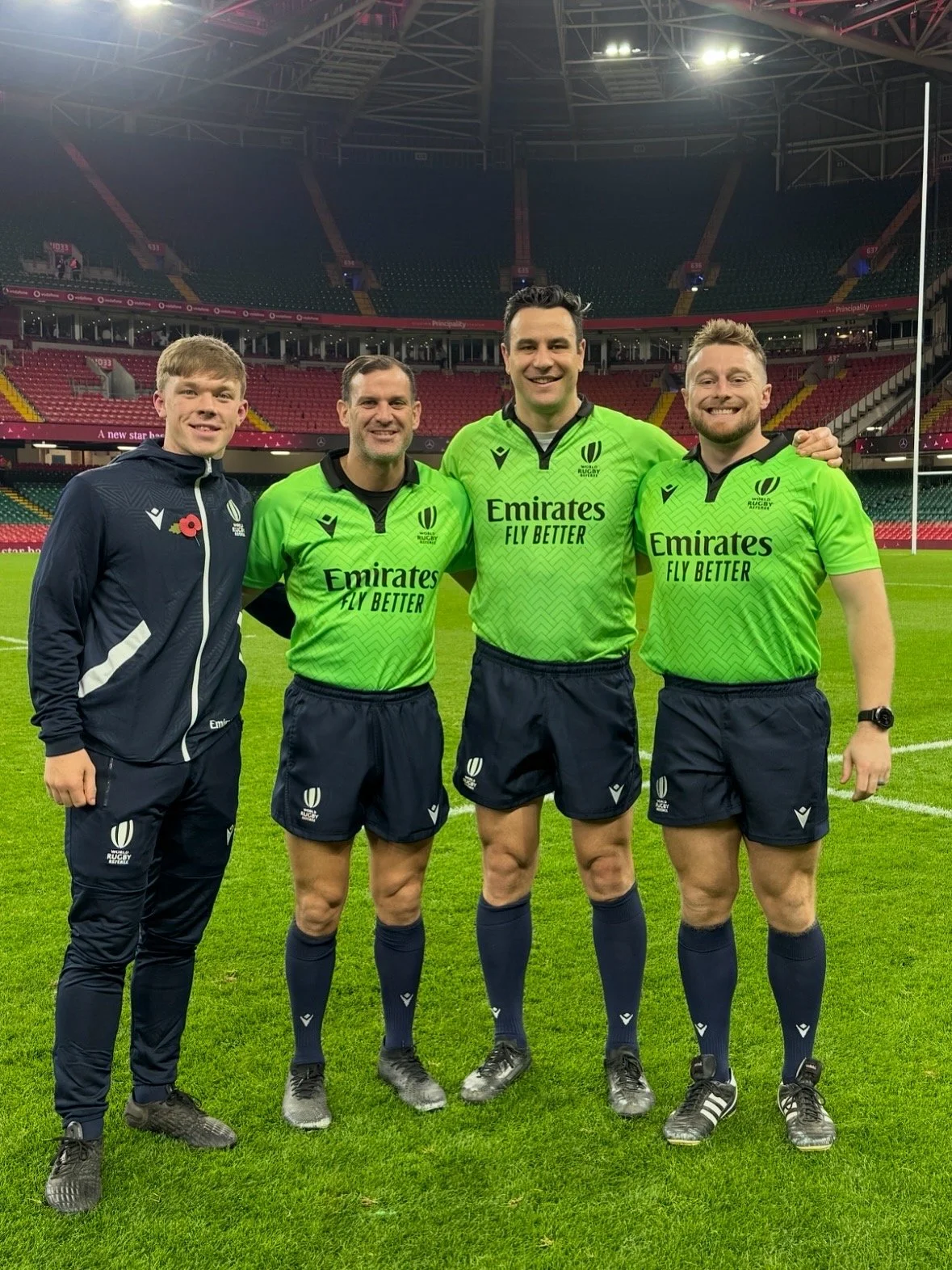 Four men standing on a rugby field inside a stadium, with three wearing bright green rugby jerseys and one in a dark jacket and pants. They are smiling and posing for the photo including Ben O'Keeffe.