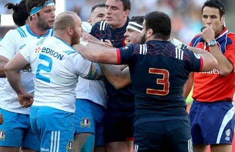 Rugby players from opposing teams engaged in a heated confrontation on the field with referee Ben O'Keeffe controlling proceedings.