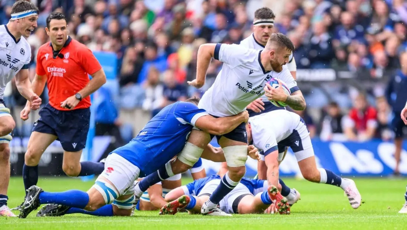 Rugby players in action during a match, with a player holding a rugby ball while being tackled by opponents, and others running or observing on the field, with a crowd in the background. Including rugby referee Ben O'Keeffe.