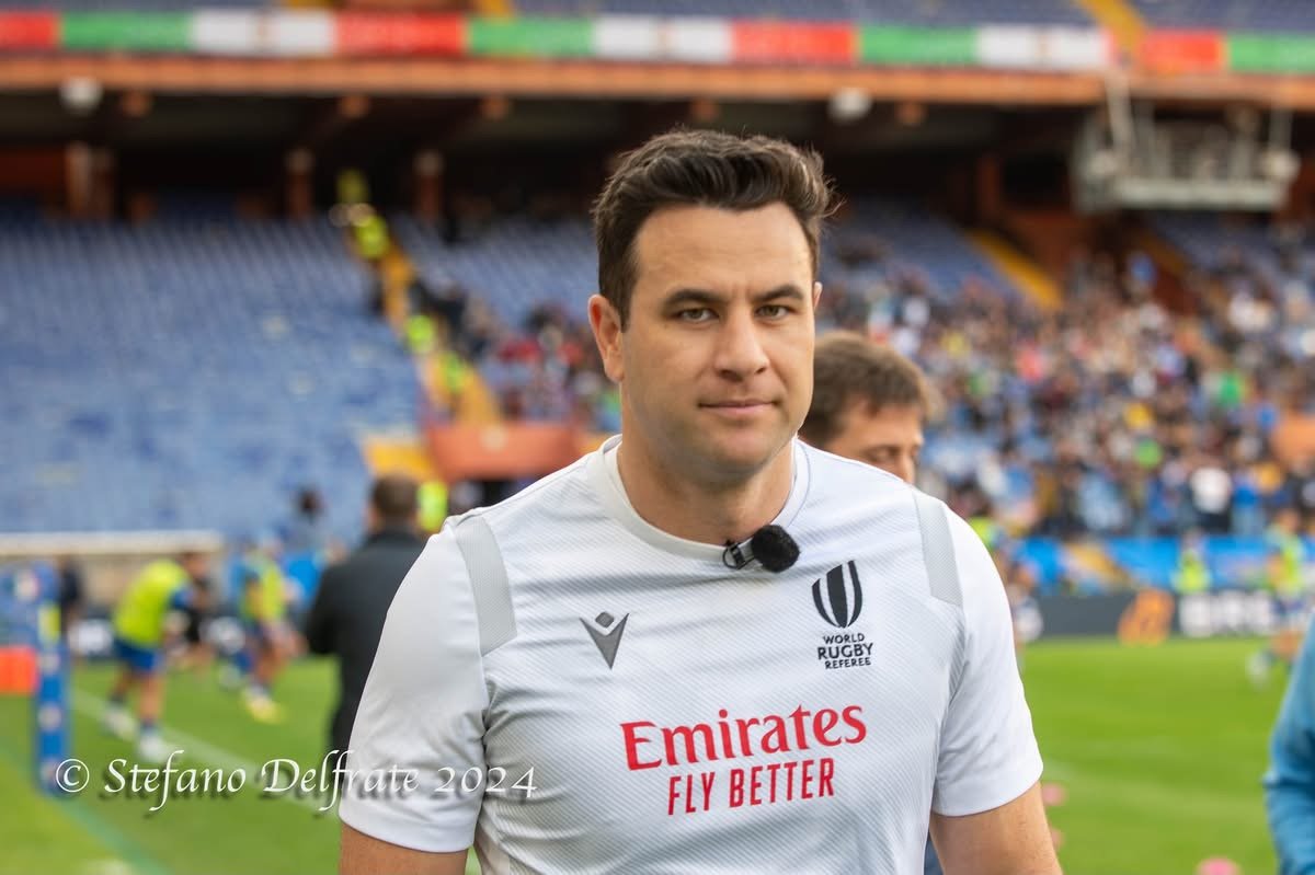 Ben O'Keeffe in a white rugby jersey with sponsor logos, standing on a rugby field with a stadium and players in the background.