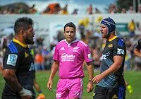 Three rugby players on the field, two wearing black and yellow uniforms and one in pink is referee Ben O'Keeffe, standing and talking during a game.