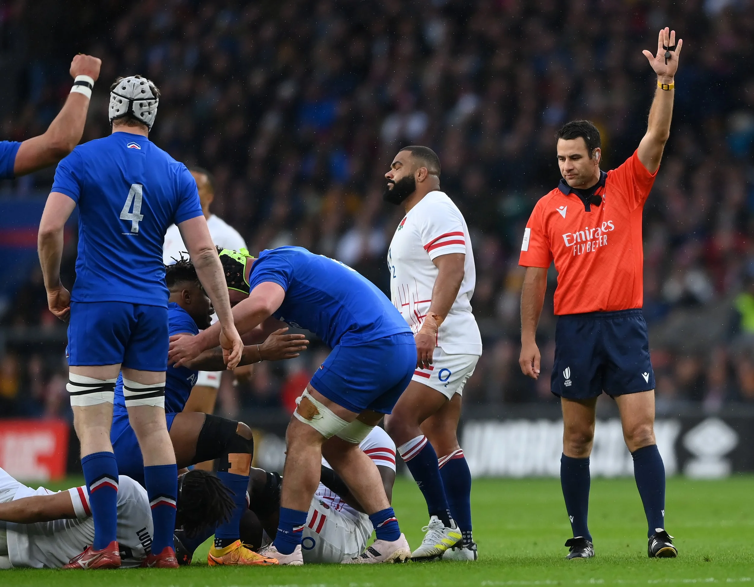 Rugby players on the field during a match, with one referee Ben O'Keeffe raising his hand.