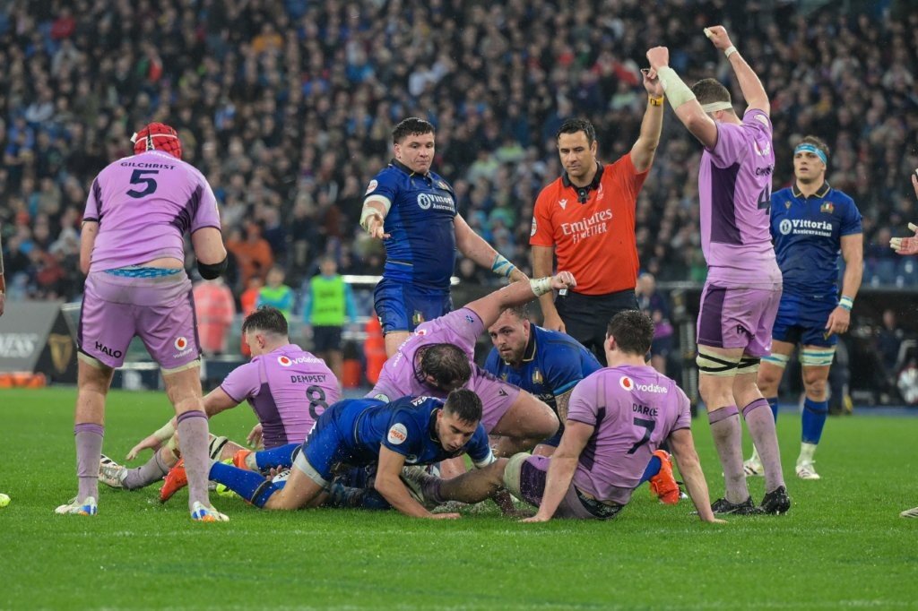 Rugby players in a match, Italy v Scotland, some on the ground and others standing around, with a referee Ben O'Keeffe in a red jersey overseeing the game, in a stadium filled with spectators.