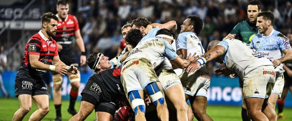 Rugby match with players engaged in a tackle and scrum on the field, some players are dirty and wearing uniforms with logos with referee Ben O'Keeffe.