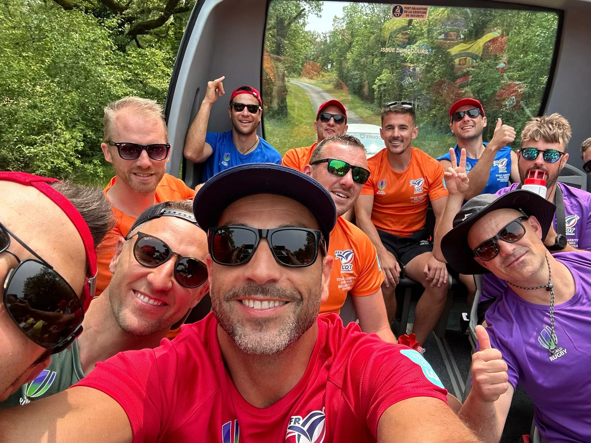 Group of smiling men on a bus or tram taking a selfie, wearing colorful sports jerseys and sunglasses, with greenery outside the window behind them including Ben O'Keeffe.