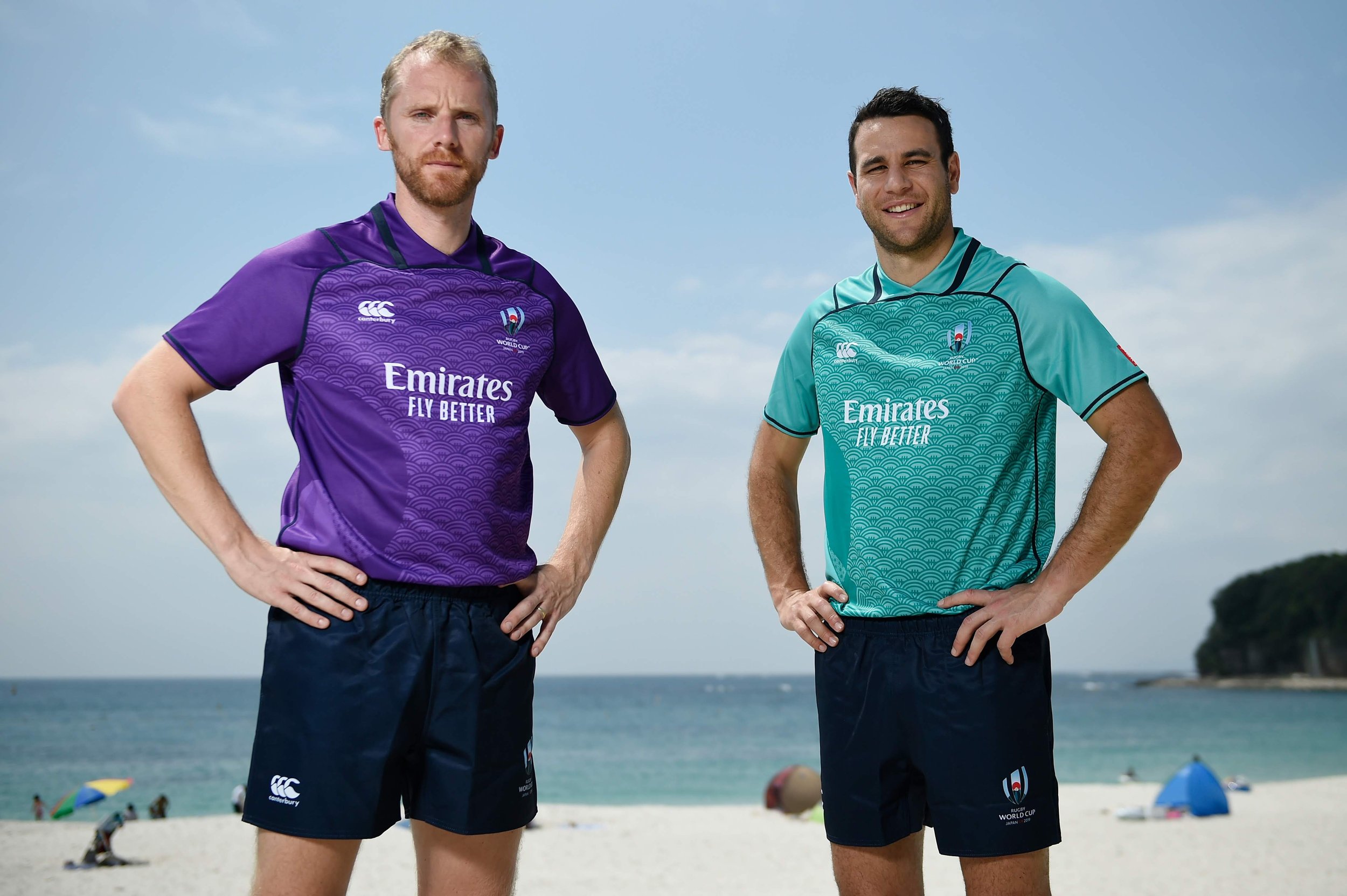 Two rugby referees standing on a beach, wearing colorful jerseys with the 'Emirates Fly Better' logo and the World Cup branding, with umbrellas and people in the background. Including Ben O'Keeffe and Wayne Barnes.