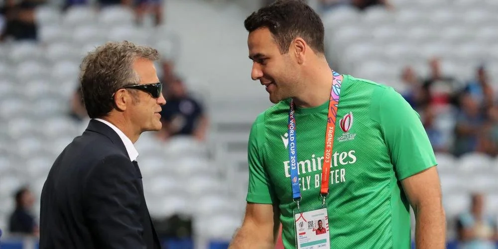 Two men in friendly conversation, one wearing a black suit and sunglasses, the other is Ben O'Keeffe in a green referee jersey with a France World Cup badge, standing on a sports stadium field.