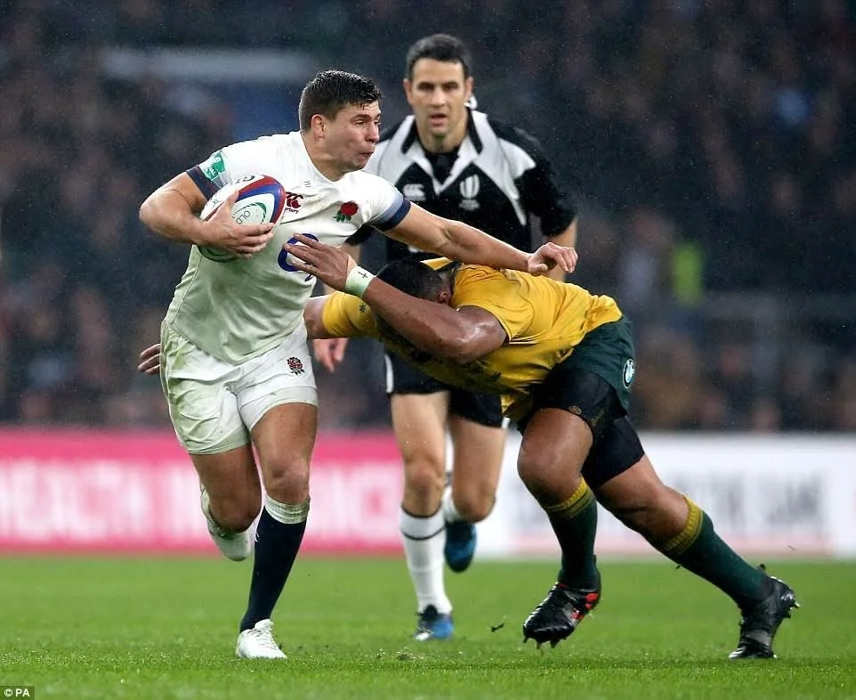 A rugby game with a player in a white uniform running with the ball while being tackled by a player in a yellow uniform. An official Ben O'Keeffe is black and white is in the background.