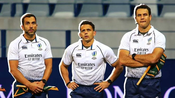 Three male rugby referees standing on the field, holding a flag, wearing white uniforms with sponsor logos. The referee is Ben O'Keeffe  during the Lions series in South Africa.