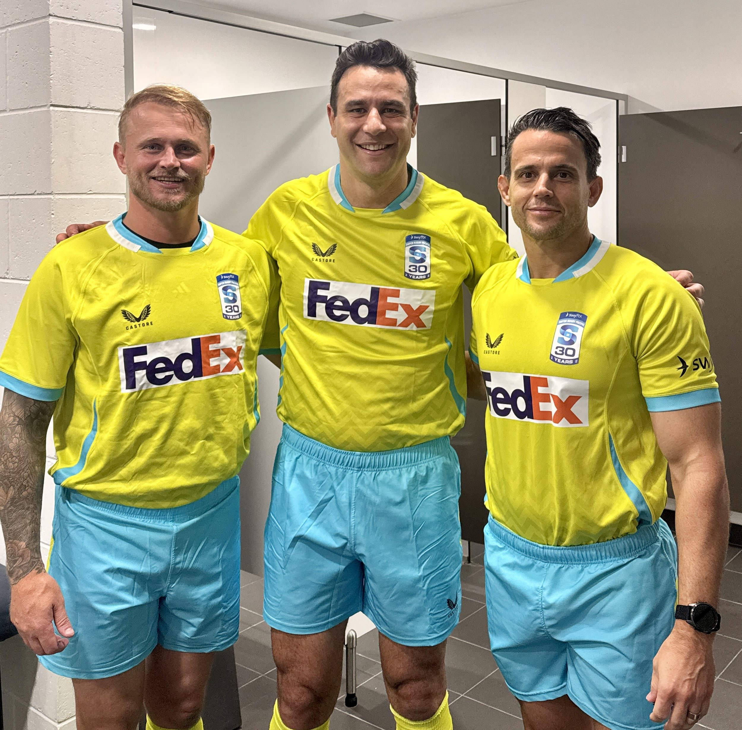 Three male referees, including Ben O'Keeffe, in yellow and turquoise sports uniforms, standing together in a locker room, smiling at the camera.