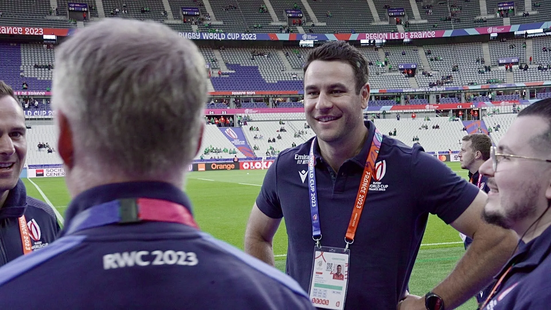 A group of officials including Ben O'Keeffe and staff on a rugby field, smiling and talking after a match at the Rugby World Cup 2023 in France, with a stadium in the background.