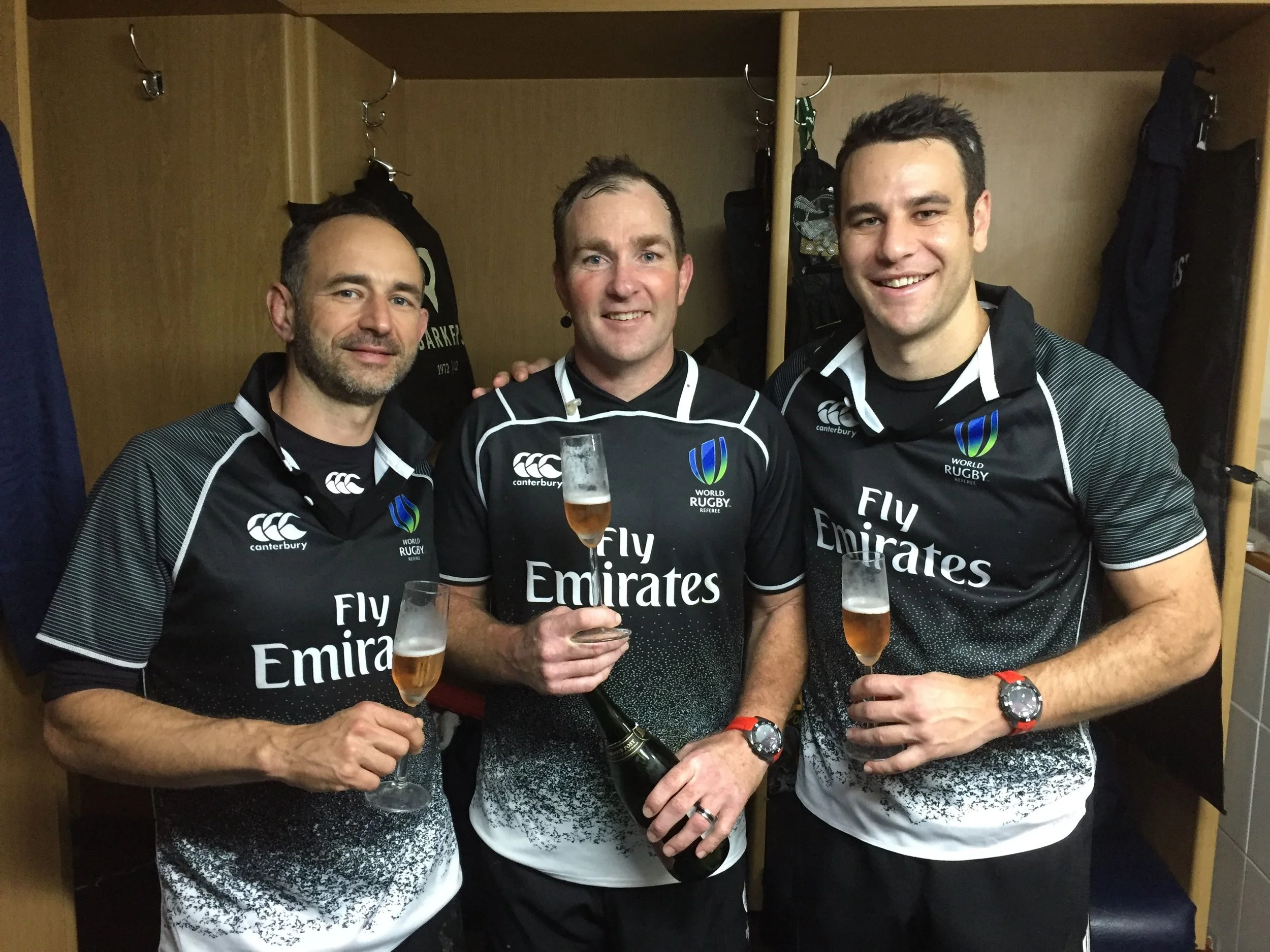 Three men in rugby referee uniforms celebrating with champagne in a locker room including Ben O'Keeffe.