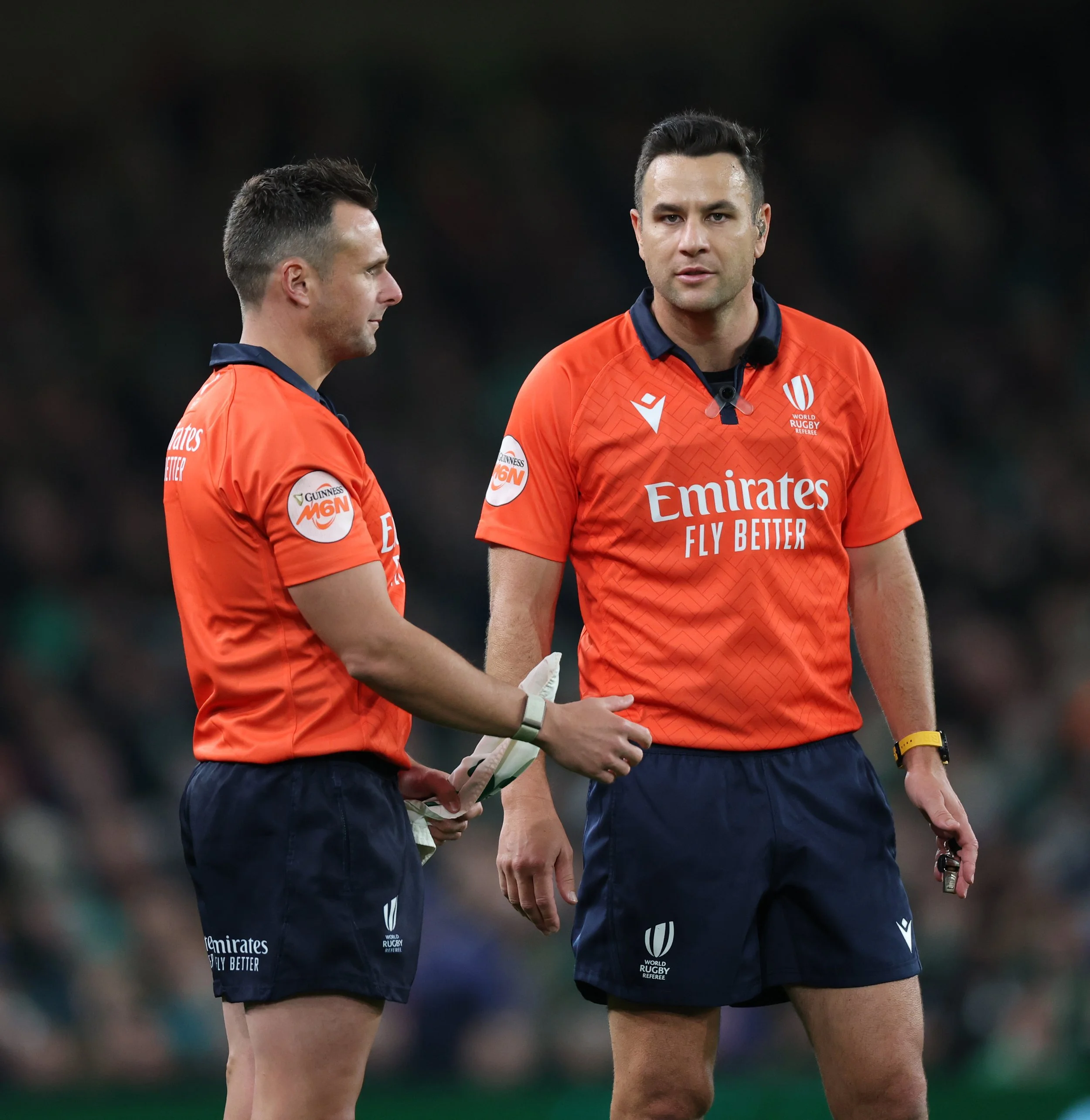 Two male rugby referees including Ben O'Keeffe in orange and navy uniforms standing on a field, one holding a piece of paper.
