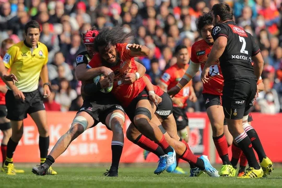 Rugby players in red and black jerseys compete for possession of the ball on the field during a game, with spectators in the stands and referee Ben O'Keeffe.