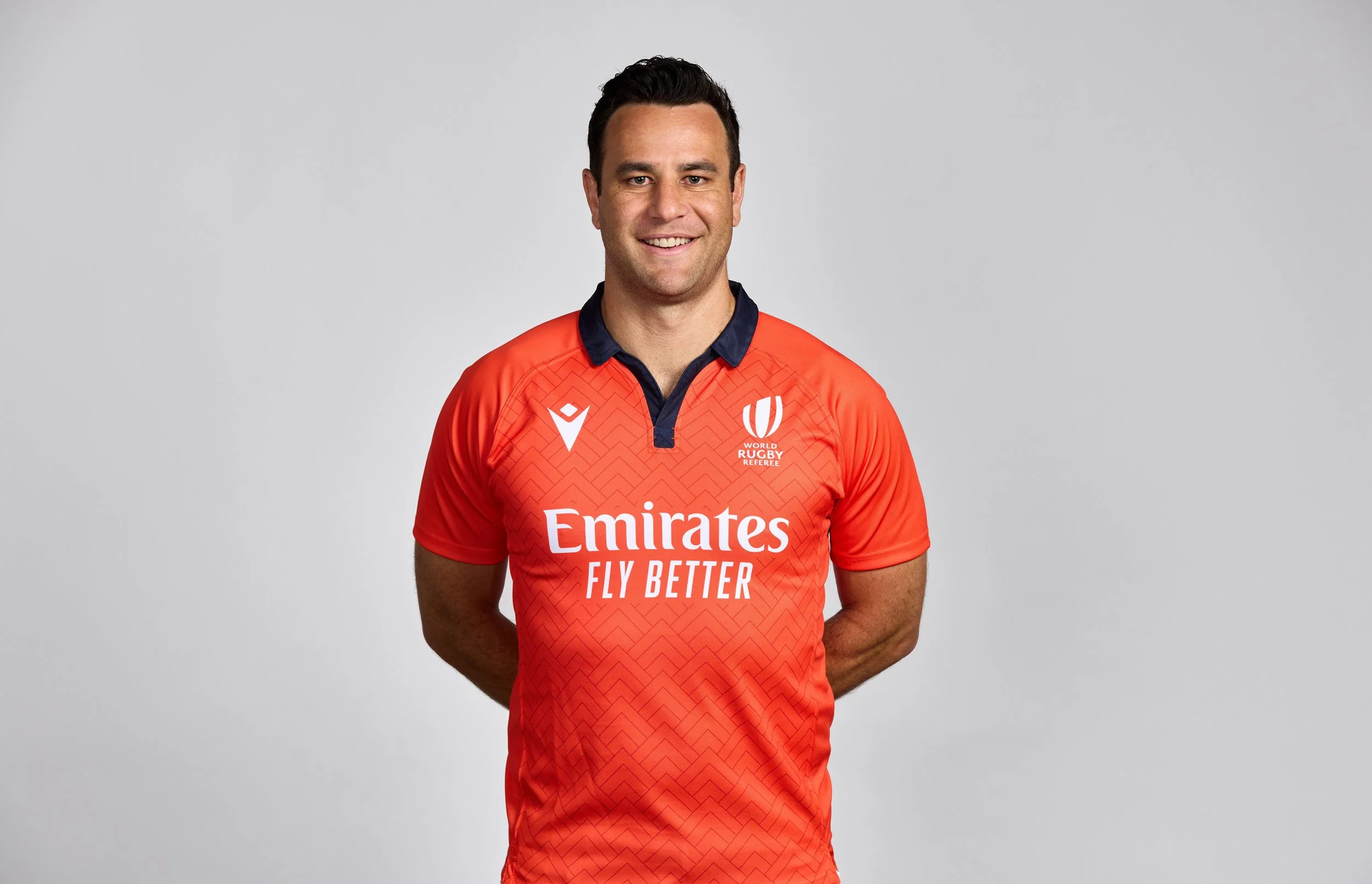 Ben O'Keeffe wearing a red rugby jersey with the logos of Emirates, World Rugby Referee, and a sports brand, standing against a plain gray background, smiling, with his hands behind his back.