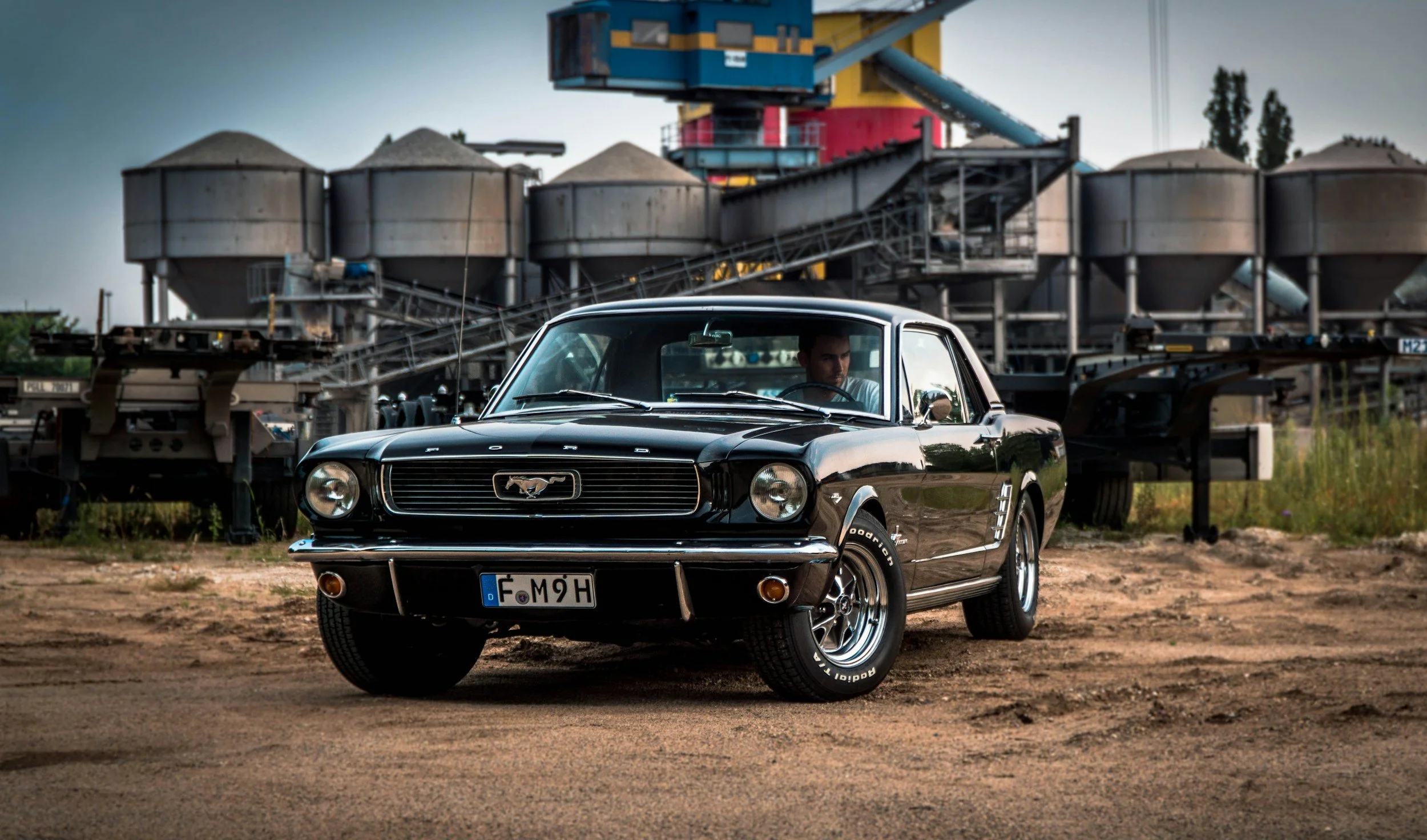 A vintage black Ford Mustang car parked on dirt ground with industrial silos and machinery in the background.
