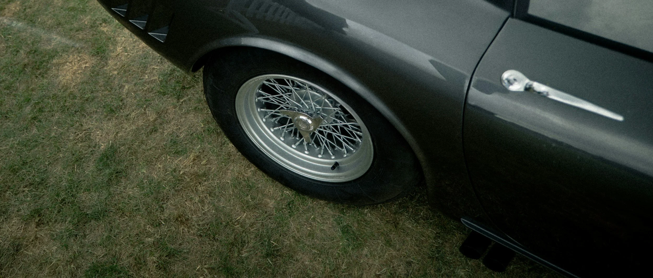 Close-up of a classic black car with chrome wire-spoke wheel and chrome door handle, parked on grass.