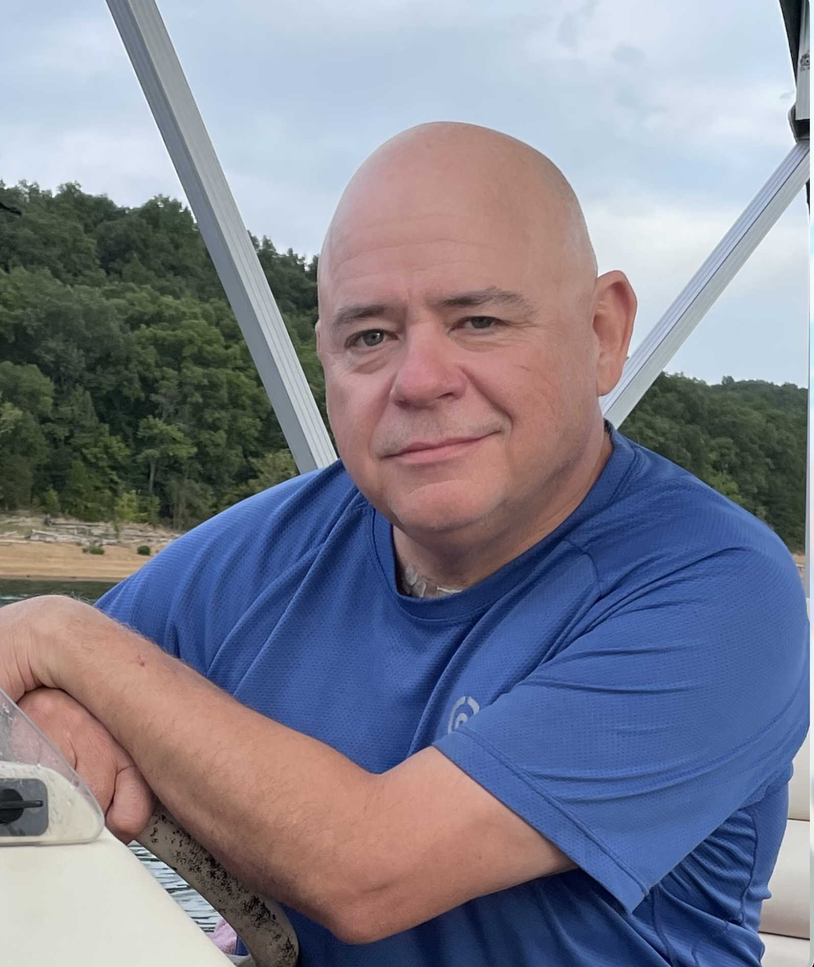 A man with a shaved head and light skin is sitting outdoors on a boat with water and green trees in the background, wearing a blue shirt.
