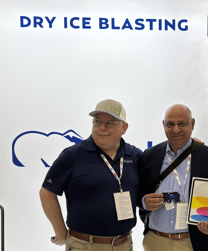 Two men standing in front of a sign that says 'Dry Ice Blasting.' One man is wearing a navy polo shirt, glasses, and a beige cap, while the other man is dressed in a suit jacket and blue dress shirt. They are smiling and holding various items, with conference badges hanging around their necks.