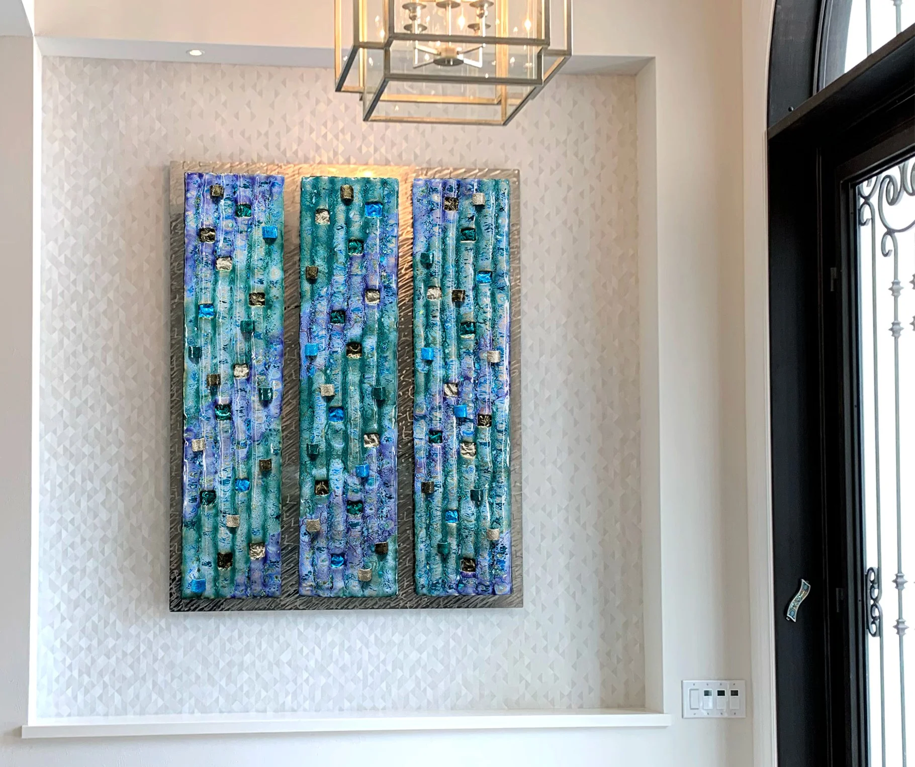 A three-panel abstract art piece in shades of blue, turquoise, and silver, mounted on a cream-colored wall with a textured pattern, beneath a modern rectangular light fixture.