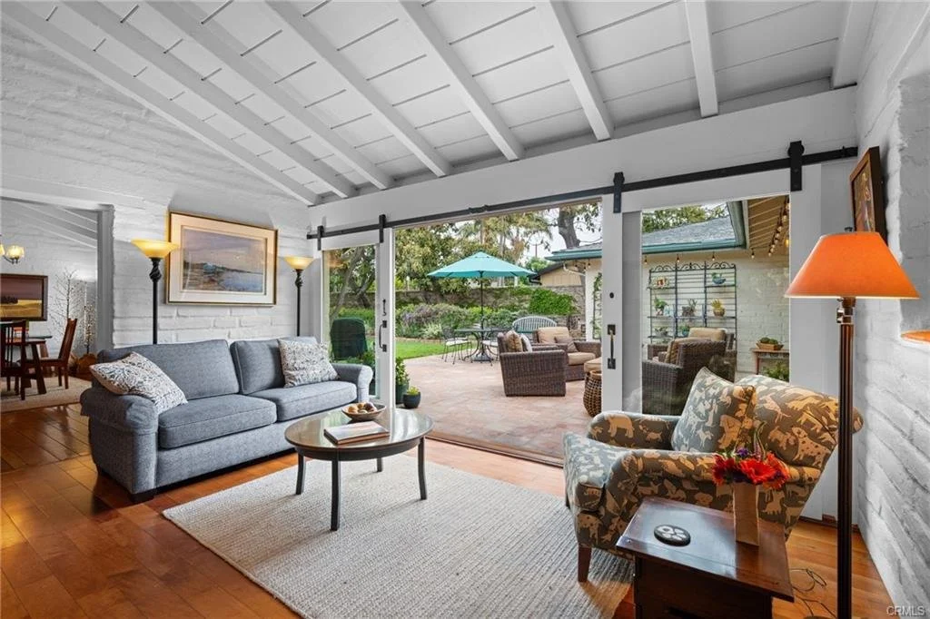 Living room with a large sliding glass door leading to a backyard patio with outdoor seating and a green umbrella. The room has a gray sofa, a patterned armchair, a small wooden side table with a lamp, and a ceiling with white paneling.