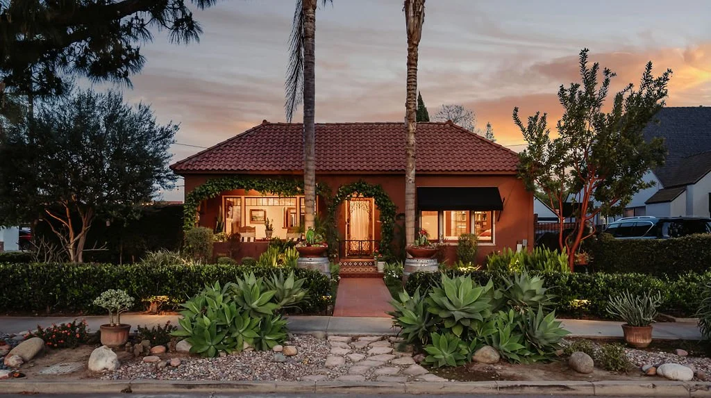 A house with a red tile roof, surrounded by a landscaped yard with plants, bushes, and trees. The house is lit from inside, and the sky shows a sunset or sunrise.