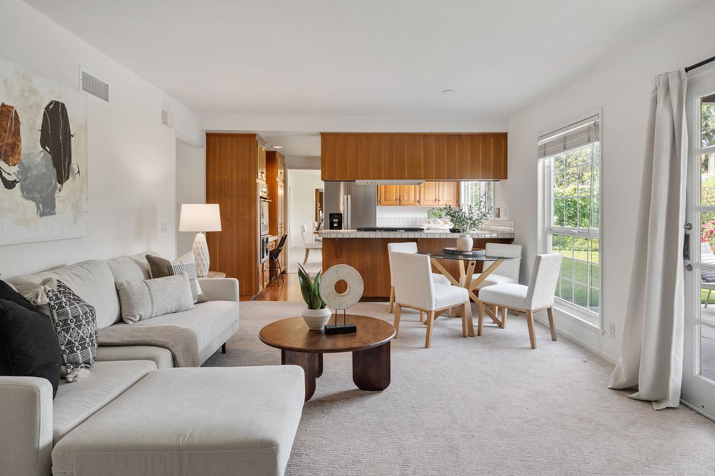 Open-concept living room and dining area with large windows, white walls, neutral-colored furniture, and a view of a green outdoor space.