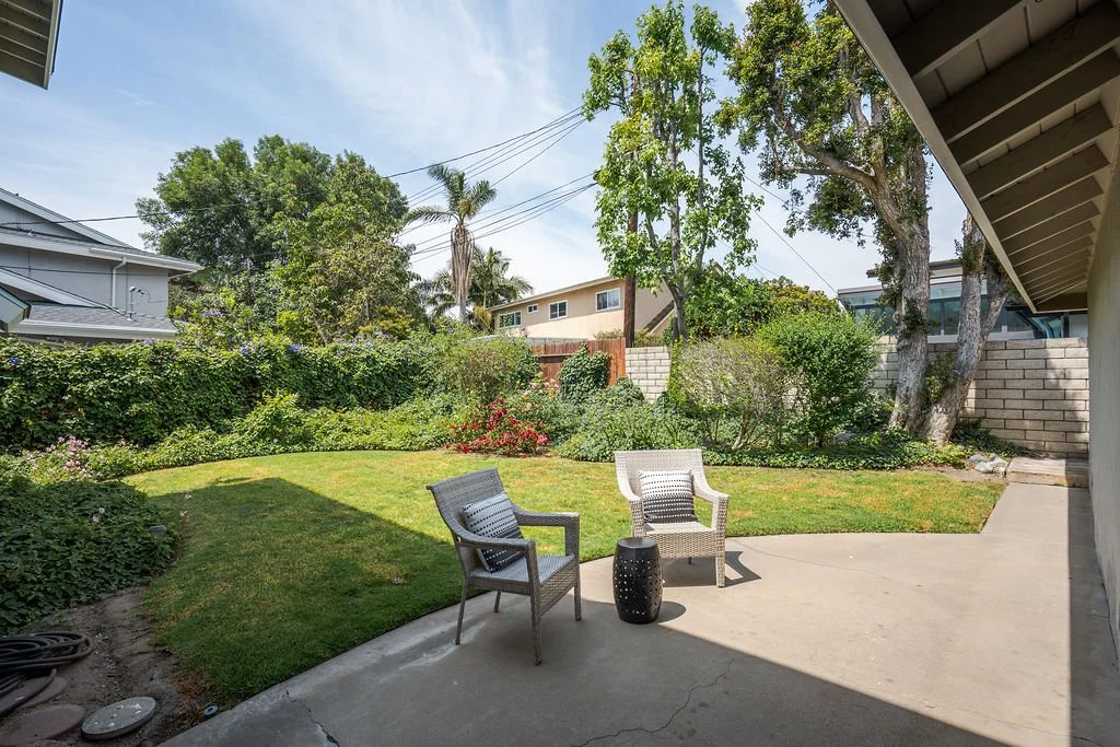 A backyard with a patio, two chairs, a small black table, a grassy lawn, trees and flowering bushes, and a wooden fence with neighboring houses in the background.