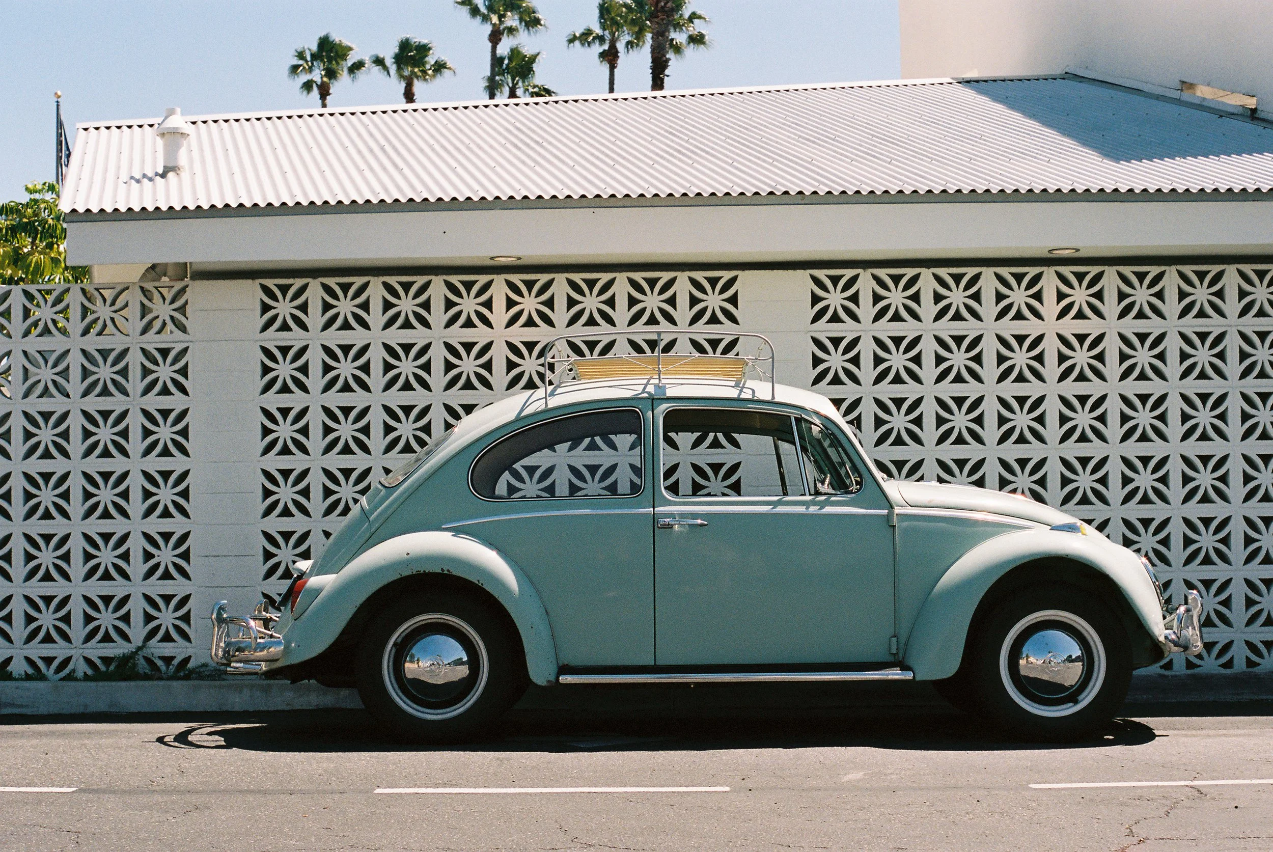 A light blue vintage Volkswagen Beetle car parked on the street in front of a white concrete wall with decorative geometric patterns and a building with a white corrugated metal roof. There are palm trees in the background under a clear sky.