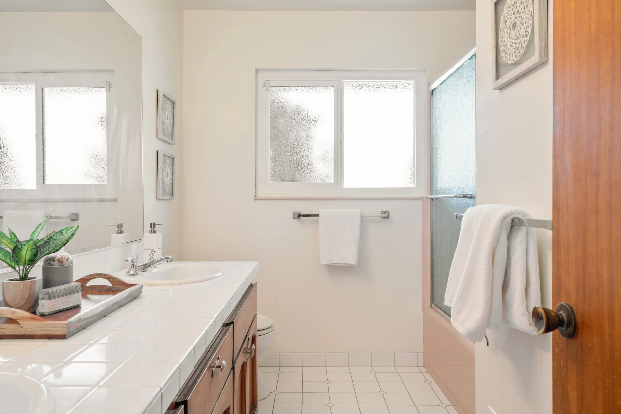 A bright bathroom with a white tiled countertop, wooden cabinets, two windows with frosted glass, a towel rack with a white towel, decorative wall art, and a partially visible shower with a frosted glass door.