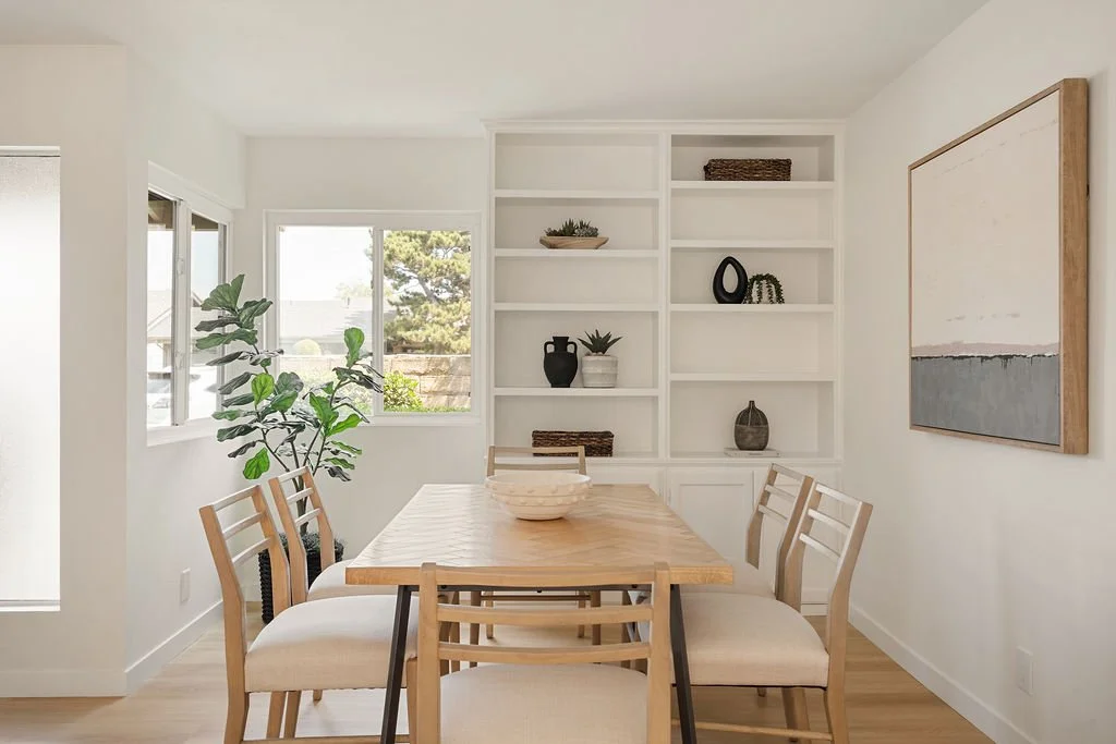 Dining room with a wooden table, six chairs with white cushions, a large white bookshelf with decorative items, a window with outside views, a potted plant, and a large abstract painting on the wall.