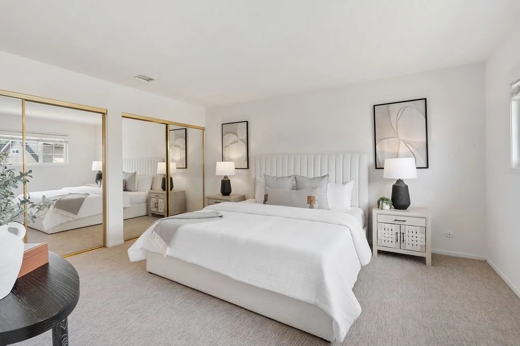 A bedroom with white walls, a white upholstered bed with multiple pillows, two nightstands with black lamps, and framed abstract artwork above the bed. There is a large closet with mirrored sliding doors and a window with blinds.