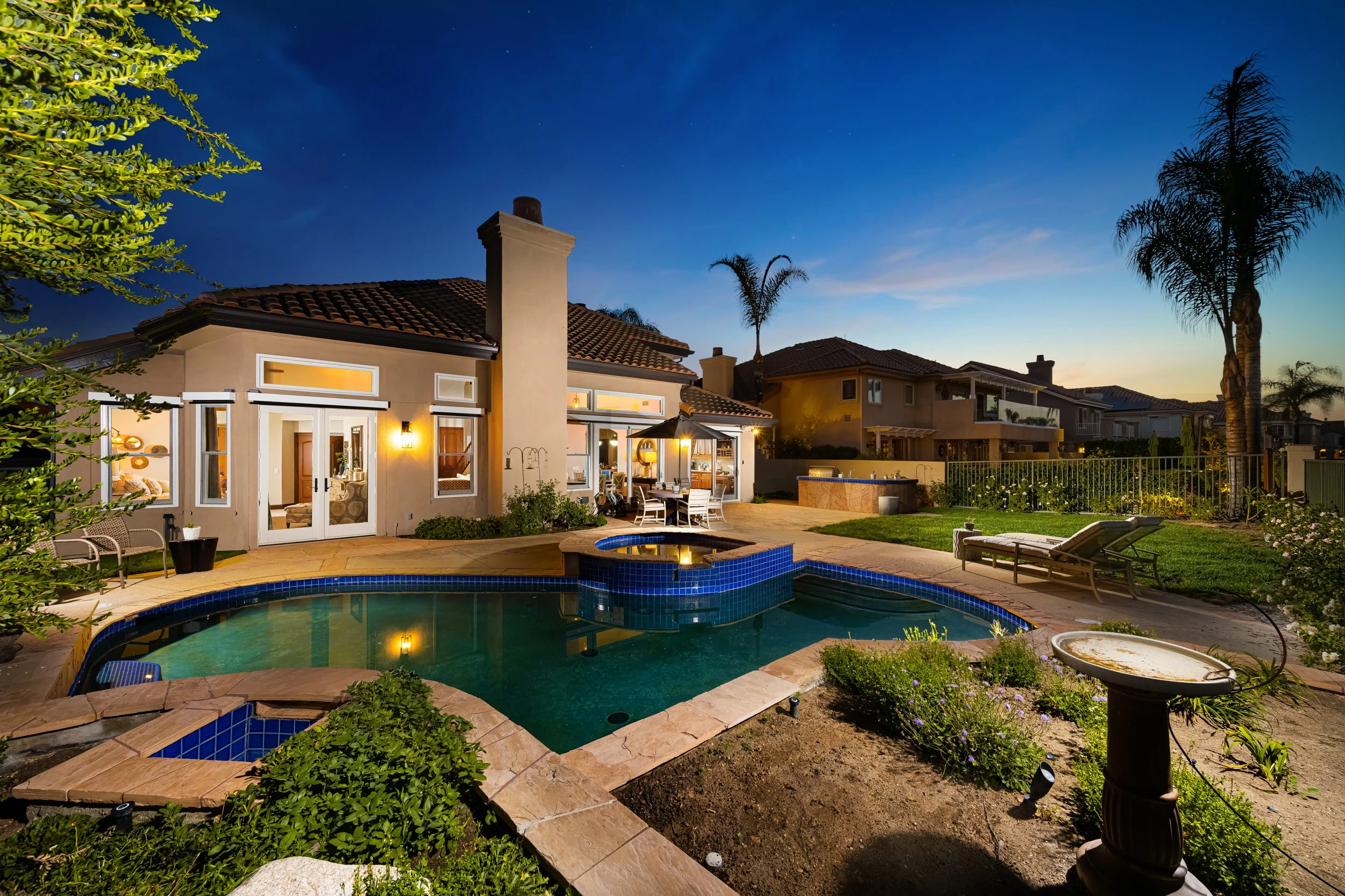 Nighttime backyard scene featuring a house with a tiled roof, lit windows, and a chimney. There is a backyard pool with blue tiles, surrounded by a stone patio, outdoor furniture, and lush greenery including palm trees and flowering plants.