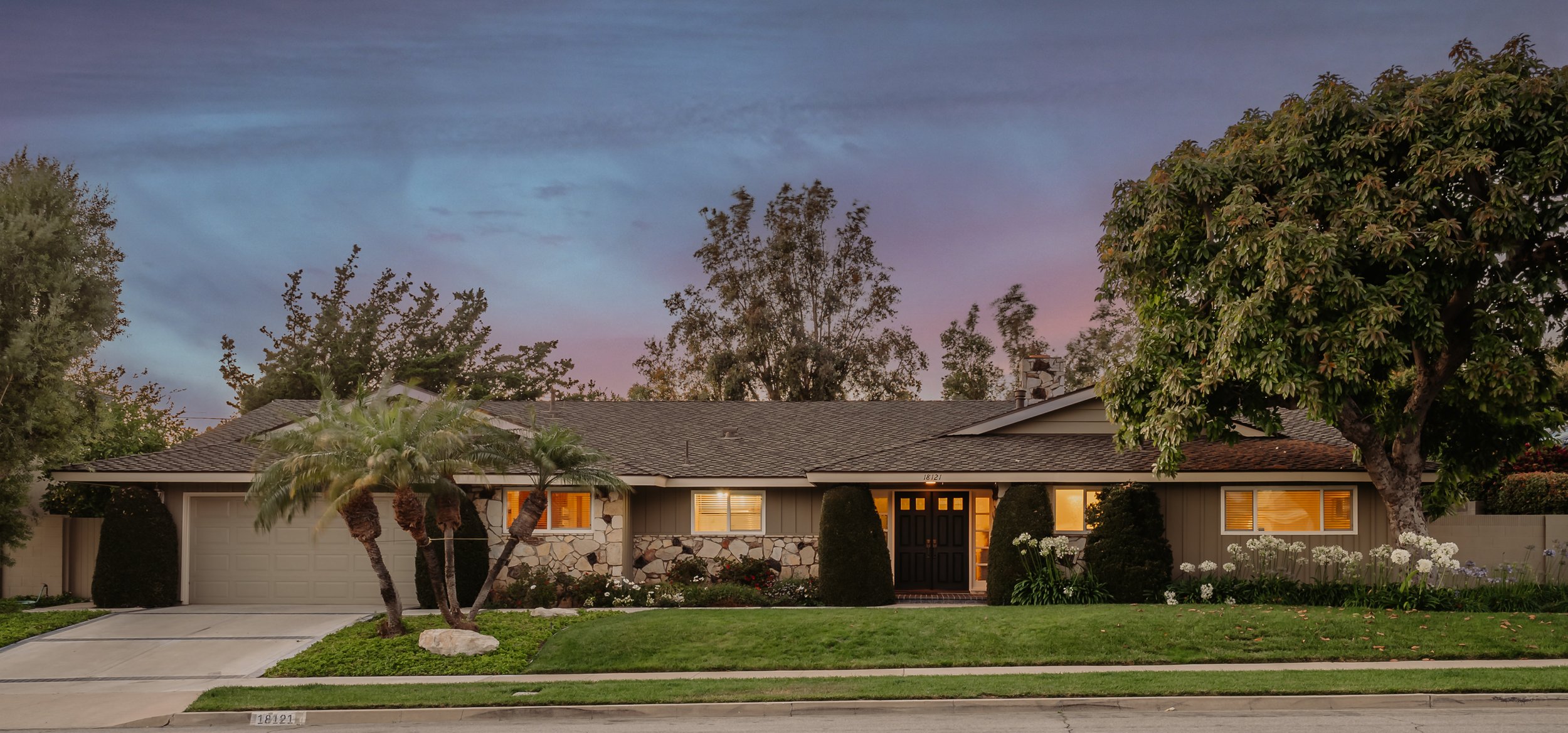 A residential house with a gray roof, stone facade, illuminated windows, and a front yard with palm trees, bushes, and white flowers, under a colorful evening sky.