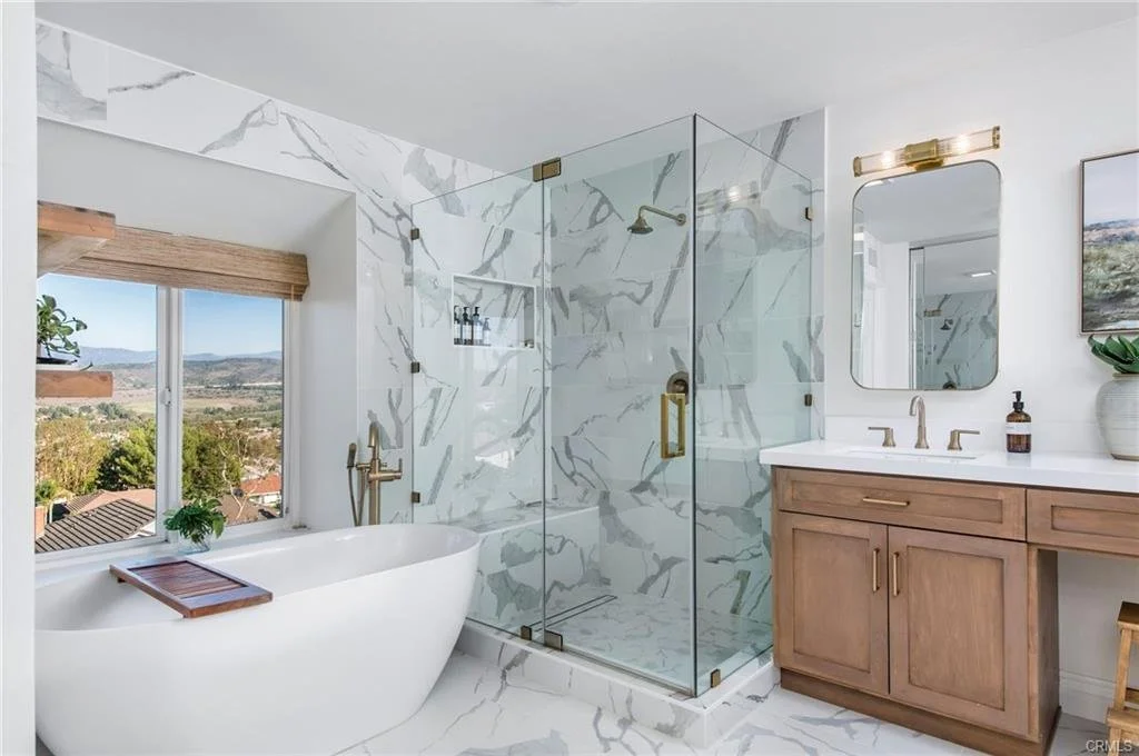 Modern bathroom with freestanding bathtub, marble walls and floors, glass shower enclosure, wooden vanity with a mirror, and a window with a scenic view.