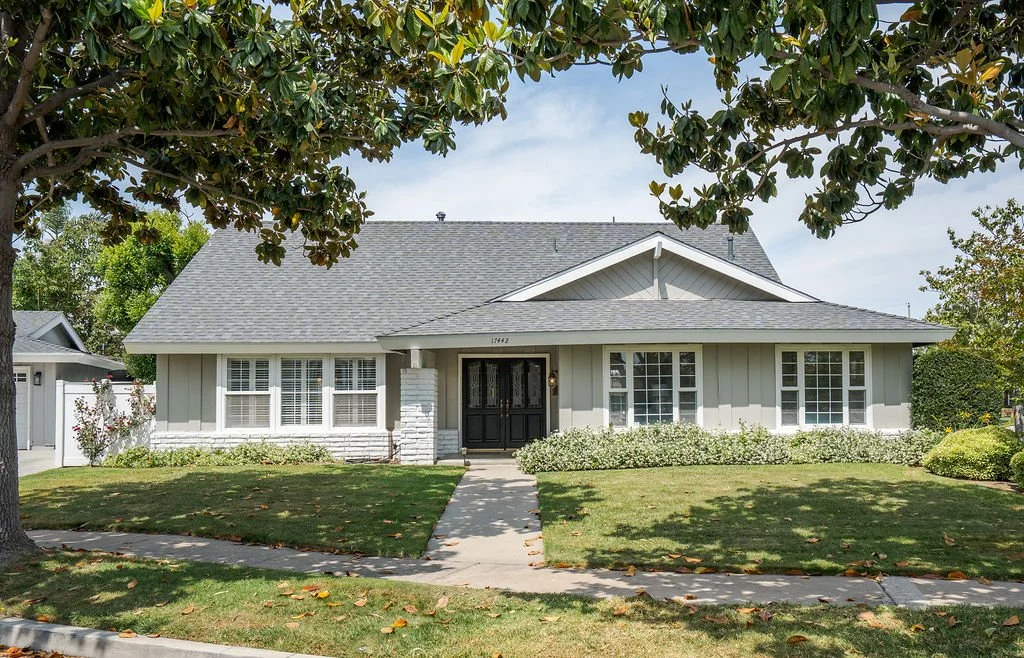 A single-story house with a grey roof and light-colored siding, surrounded by a well-maintained lawn and shrubbery, with trees in the front yard.
