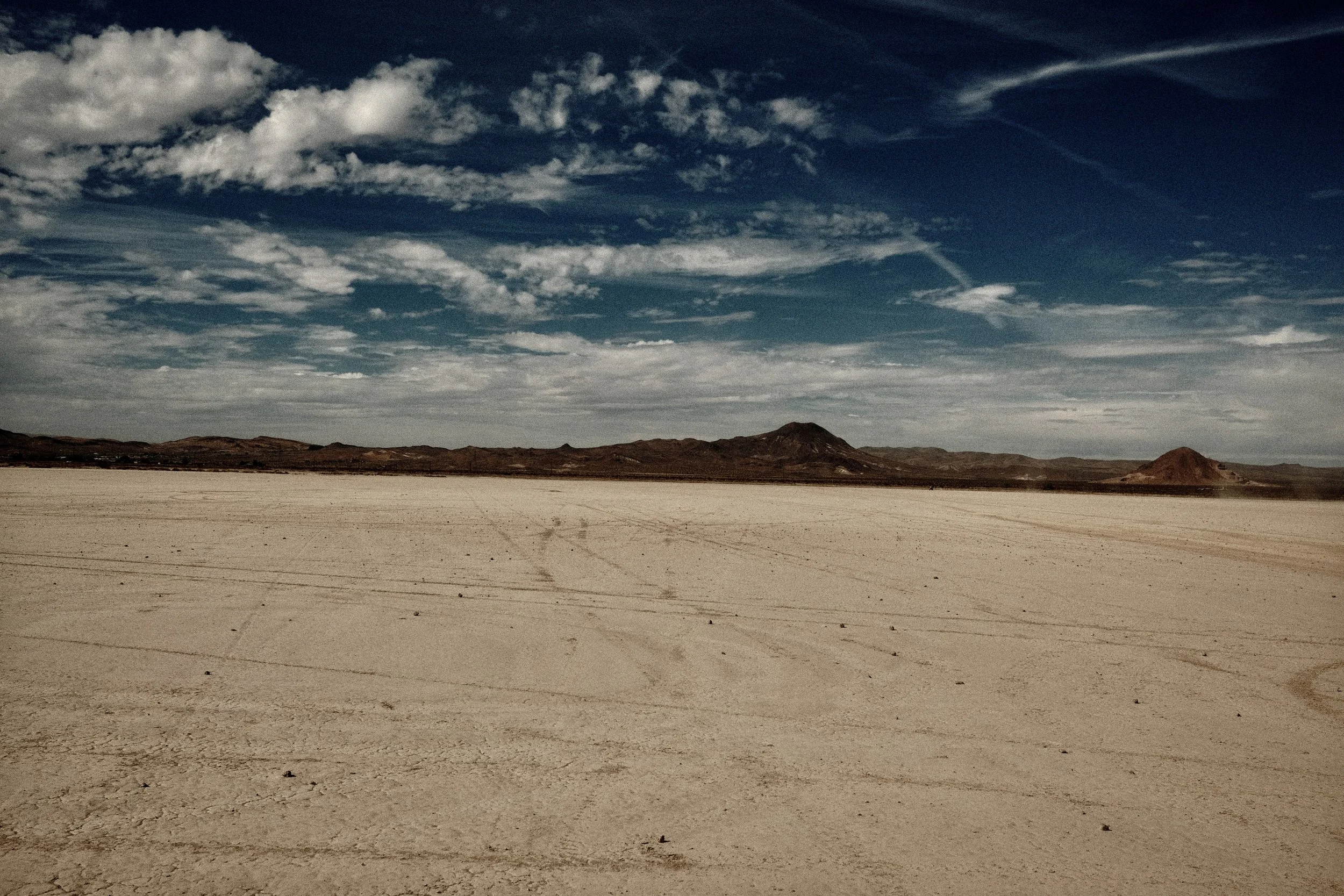 A vast, flat desert landscape with cracked white ground, distant brown hills and mountains under a blue sky with scattered clouds.