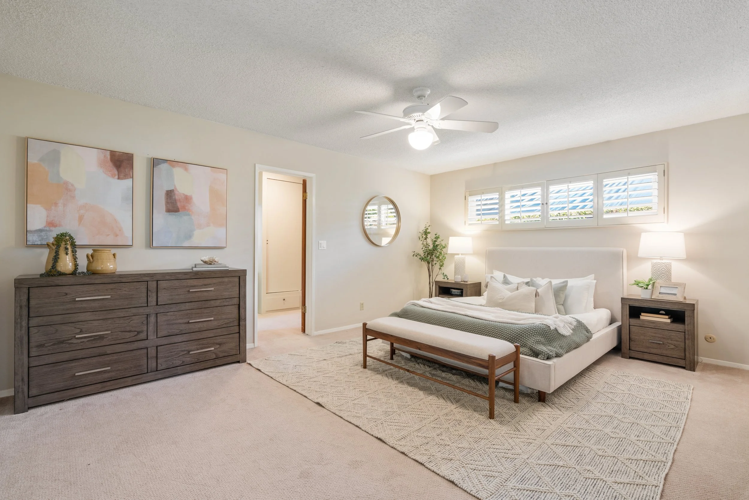 A bedroom with a white bed, two nightstands with lamps, a dresser with decorative vases, artwork on the wall, a round mirror, and a window with shutters, in a modern decor style.