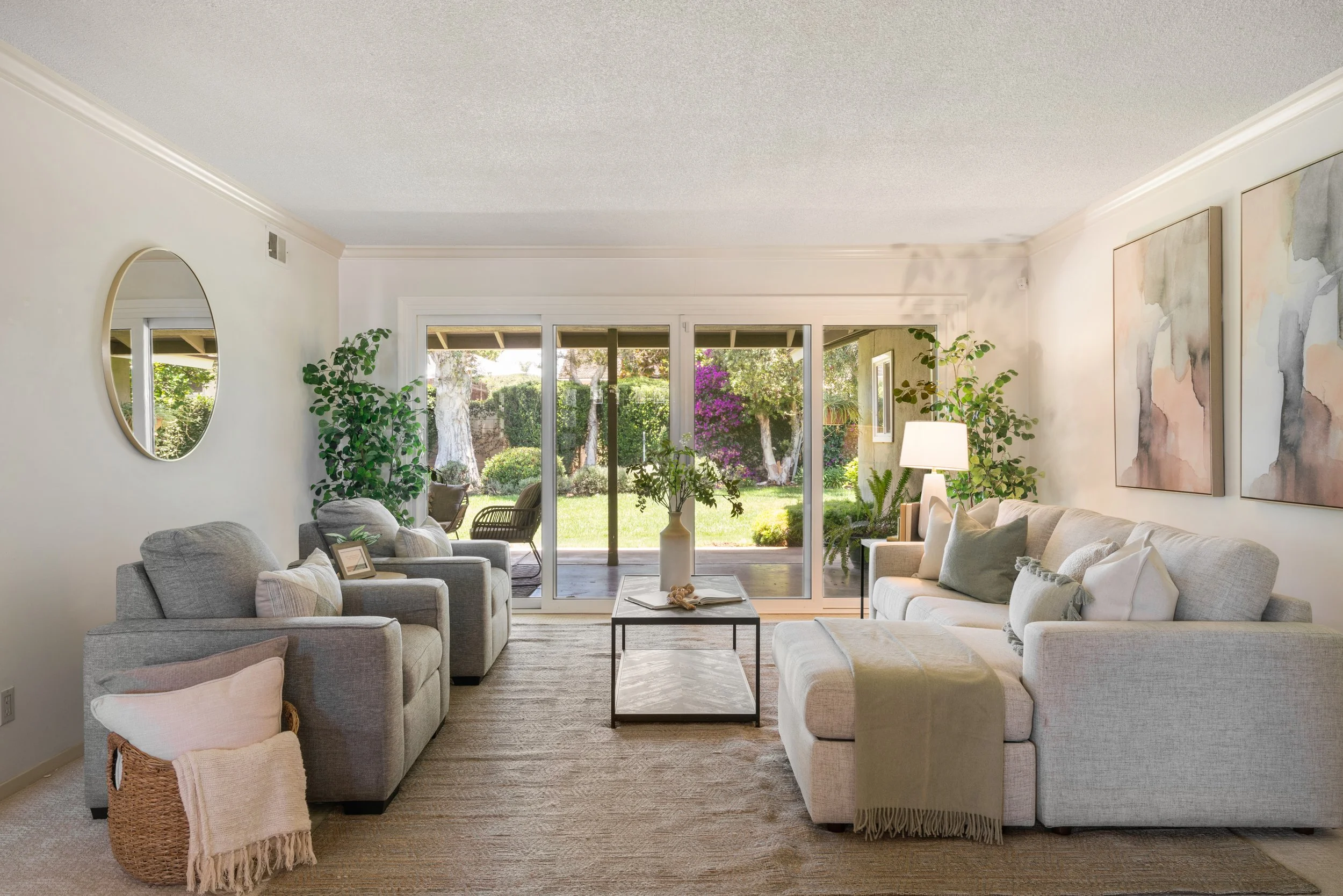 Living room with beige sofas, a coffee table, plants, artwork, and large sliding glass doors leading to a garden