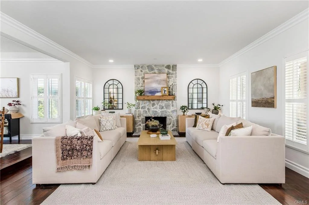Bright living room with two beige sofas, a stone fireplace with a wooden mantel, two arched black window frames, and modern decor including artwork, plants, and a wooden coffee table.