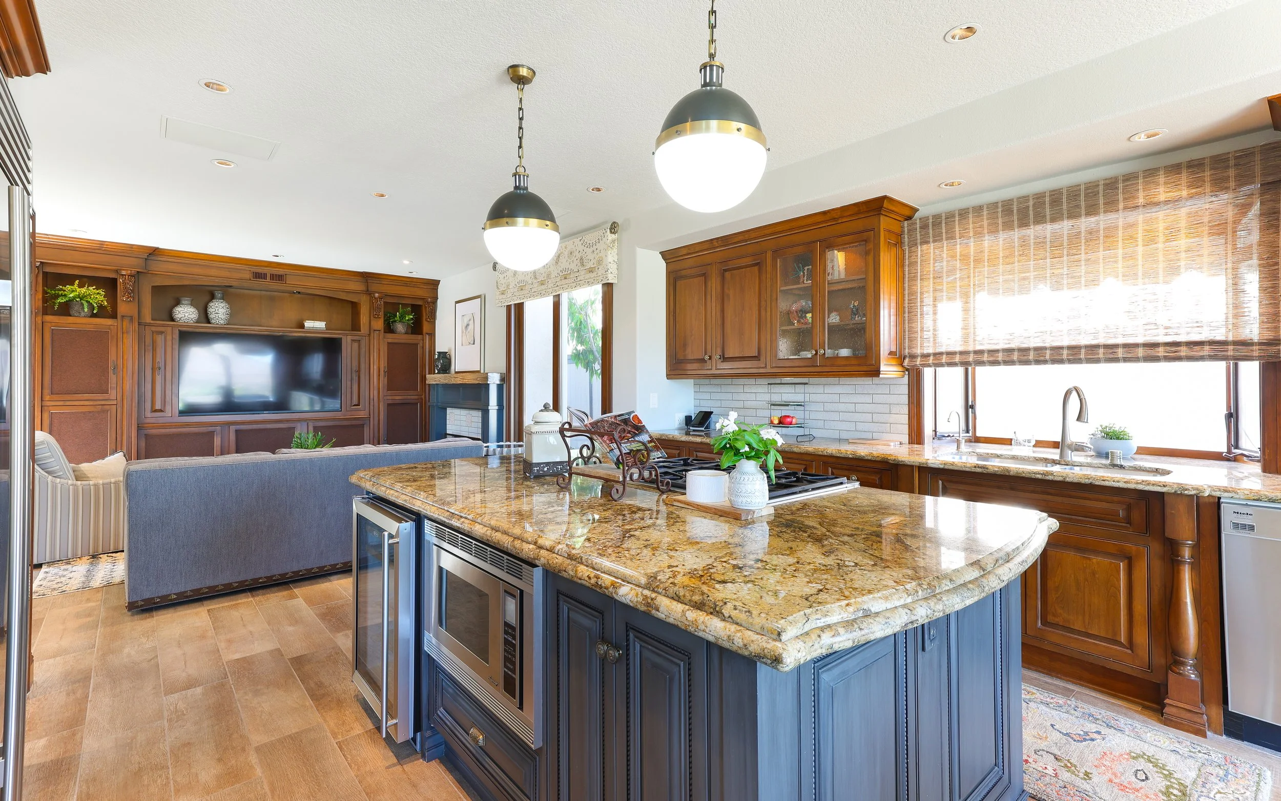A spacious open-concept kitchen and living room with wooden cabinets, granite countertops, and pendant lighting. The living area includes a TV, sofa, and decorative vases, while the kitchen features plants, a stove, and a window with bamboo blinds.