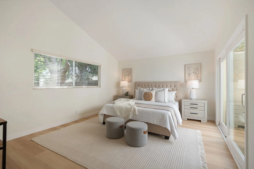 A bright, minimalist bedroom with a neutral color palette, featuring a bed with white linens, a tufted beige headboard, two nightstands with lamps, a large window with blinds, a sliding glass door, and two gray ottomans at the foot of the bed.