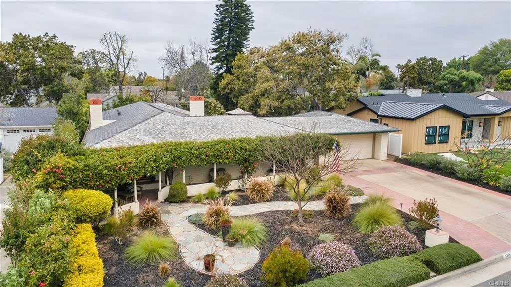 A suburban house with a spacious front yard, landscaped with a pathway, various plants, shrubs, and trees, and a driveway leading to a garage.