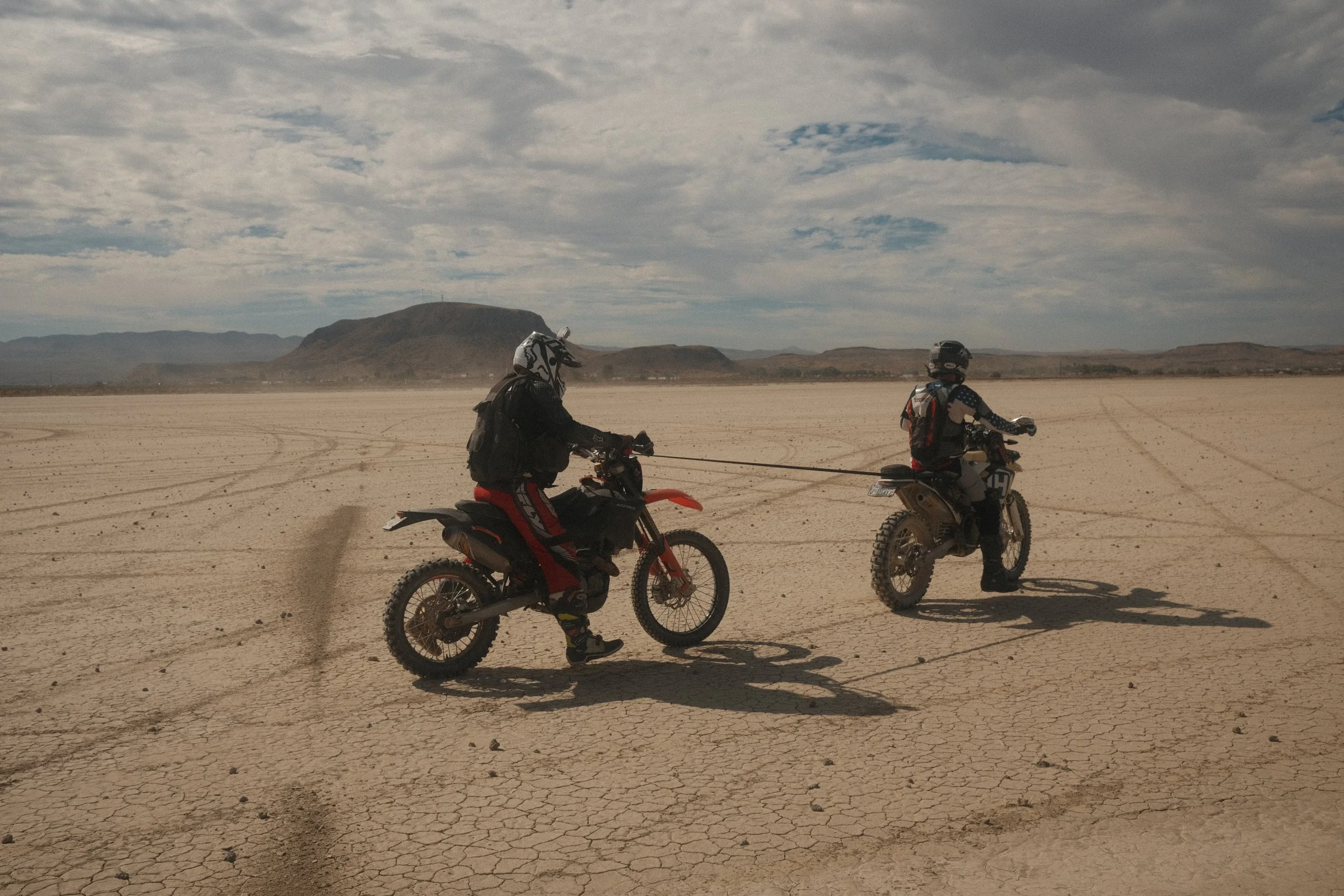 Two people riding dirt bikes in a desert landscape with mountains in the background under a cloudy sky.