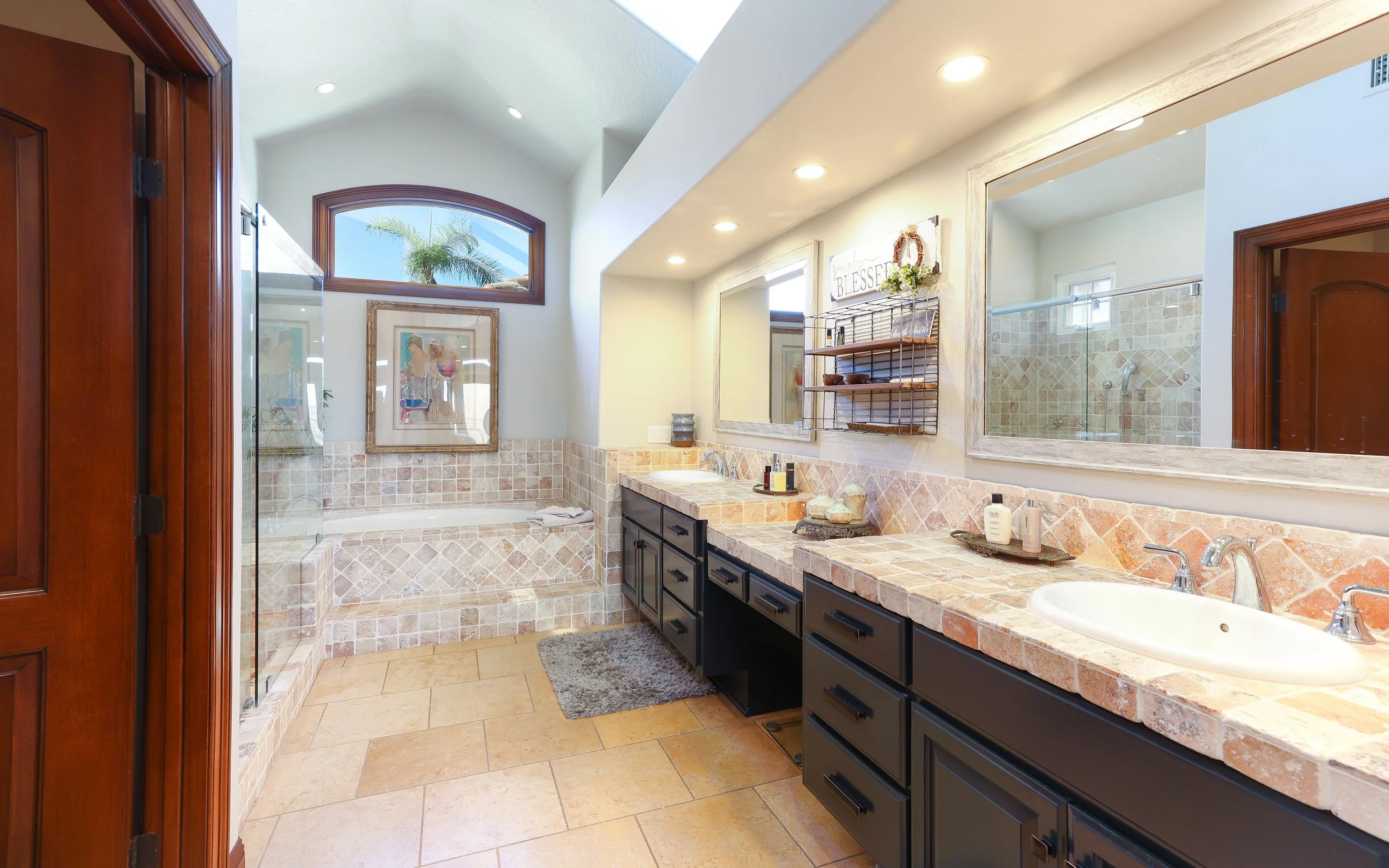 A spacious bathroom with a double sink vanity, black cabinets, and a large mirror. There is natural light coming from a window and skylight, with a decorative tile pattern on the counter and wall, and a separate shower area with glass enclosure and beige tile accents.